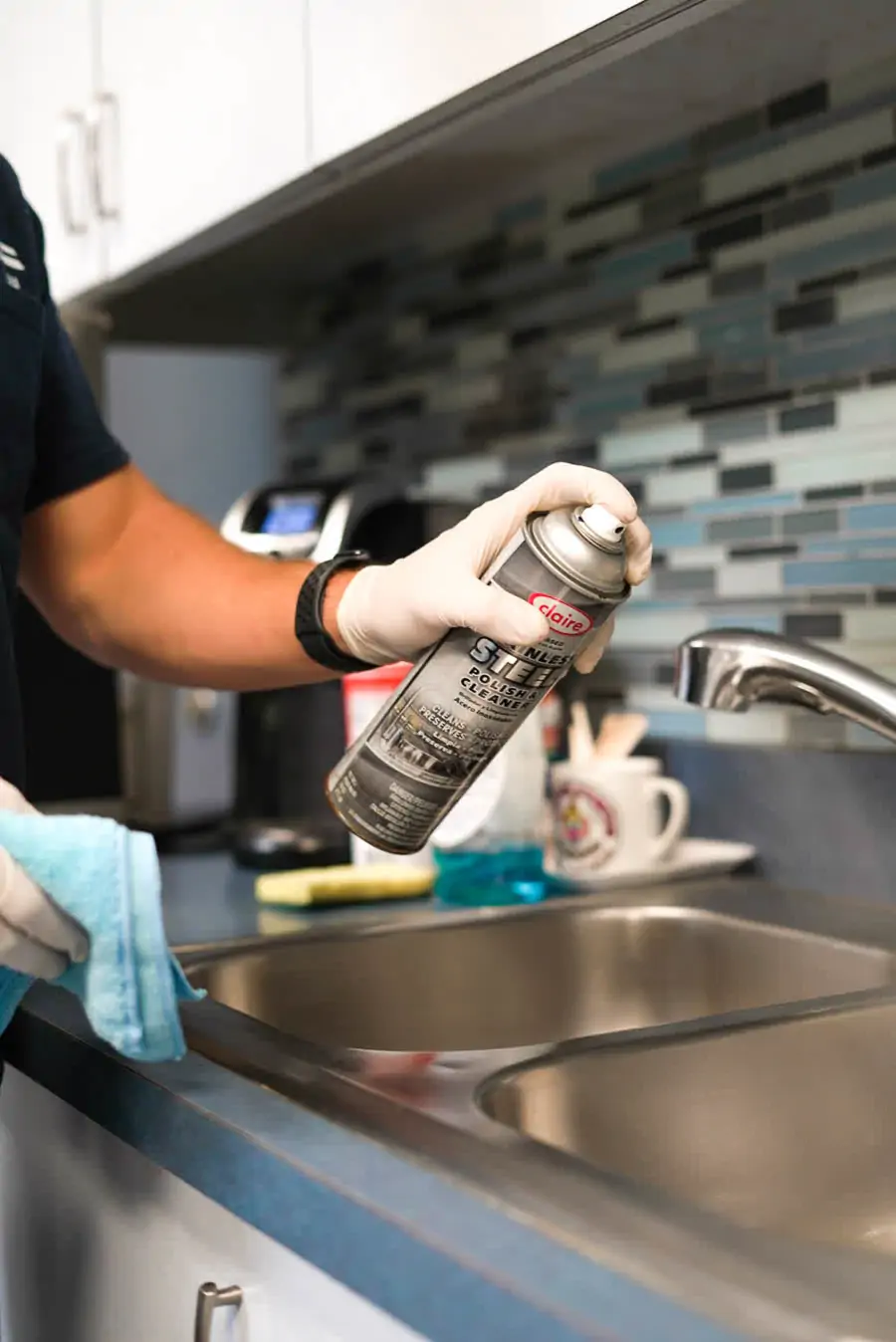 Gloved hand holding a stainless steel spray cleaner beside a commercial break room sink